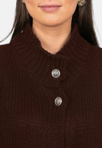 Pull en laine brun avec un col montant et deux boutons décoratifs argentés au niveau de l'encolure. La texture semble douce et texturée.