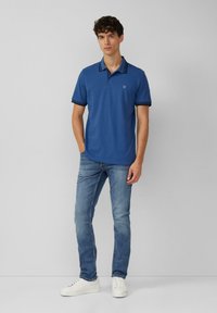 Niebieska koszulka polo z kontrastowym kołnierzykiem, krótkimi rękawami i logo na klatce piersiowej, w połączeniu z jasnymi dżinsami i białymi sneakersami.