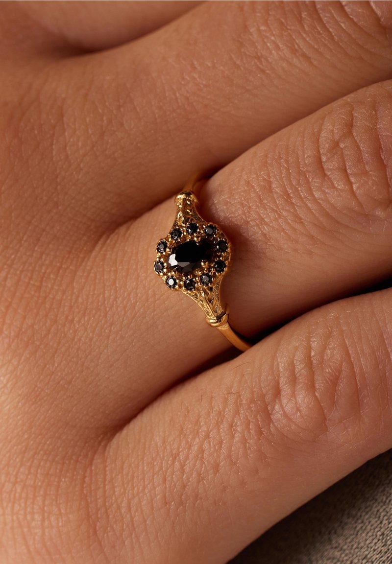 Bague en or avec une pierre précieuse noire ovale entourée de petites pierres noires sur le doigt d'une personne avec une texture de peau visible.