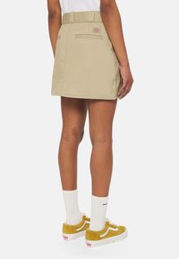 Gonna beige con vita aderente e tasche posteriori, abbinata a sneakers in suede gialle e calze bianche, che mostrano un design casual e moderno.
