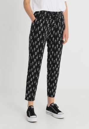 Pantaloni neri a fantasia con disegni floreali bianchi, vita elastica, tasche laterali e taglio tapered, abbinati a sneakers nere.