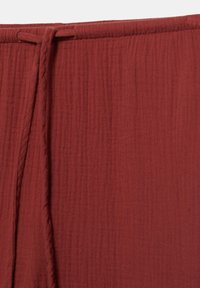 Gros plan d'un tissu texturé couleur rouille avec une sangle étroite passée dans une couture horizontale en haut.