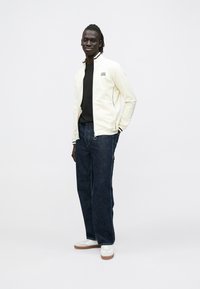 Hvit genser med glidelås og svarte detaljer, svart skjorte under, blå denimjeans og hvite joggesko med brune såler. Stående positur.