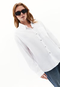 Camicia bianca a maniche lunghe con bottoni sul davanti, dotata di tasca, realizzata in tessuto leggero, abbinata a occhiali da sole neri e jeans blu.