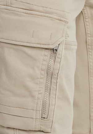 Beige Cargo-Hosen verfügen über eine mit Reißverschluss verschlossene Seitentasche, gestickte Details und einen weichen, strukturierten Stoff mit lockerer Passform.