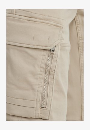 Los pantalones cargo beige cuentan con un bolsillo lateral con cremallera, detalles de costura y una tela suave y texturada con un ajuste relajado.
