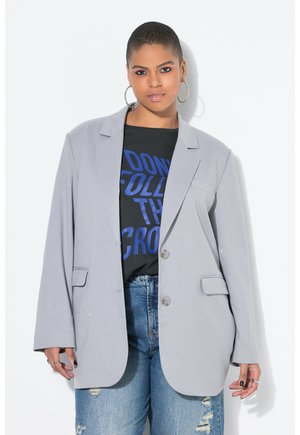 Person med kort hår iført øreringe i ringe, en lysegrå blazer over en sort skjorte med blå tekst og revnede blå jeans.