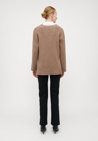 Brauner Strickpullover mit langen Ärmeln und lockerer Passform, mit geradem Saum und subtiler Textur, kombiniert mit schwarzen, taillierten Hosen.