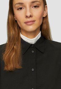 Zwart shirt met een gestructureerde afwerking, voorzien van een klassieke kraag en knopen. Een wit coltrui is eronder zichtbaar.