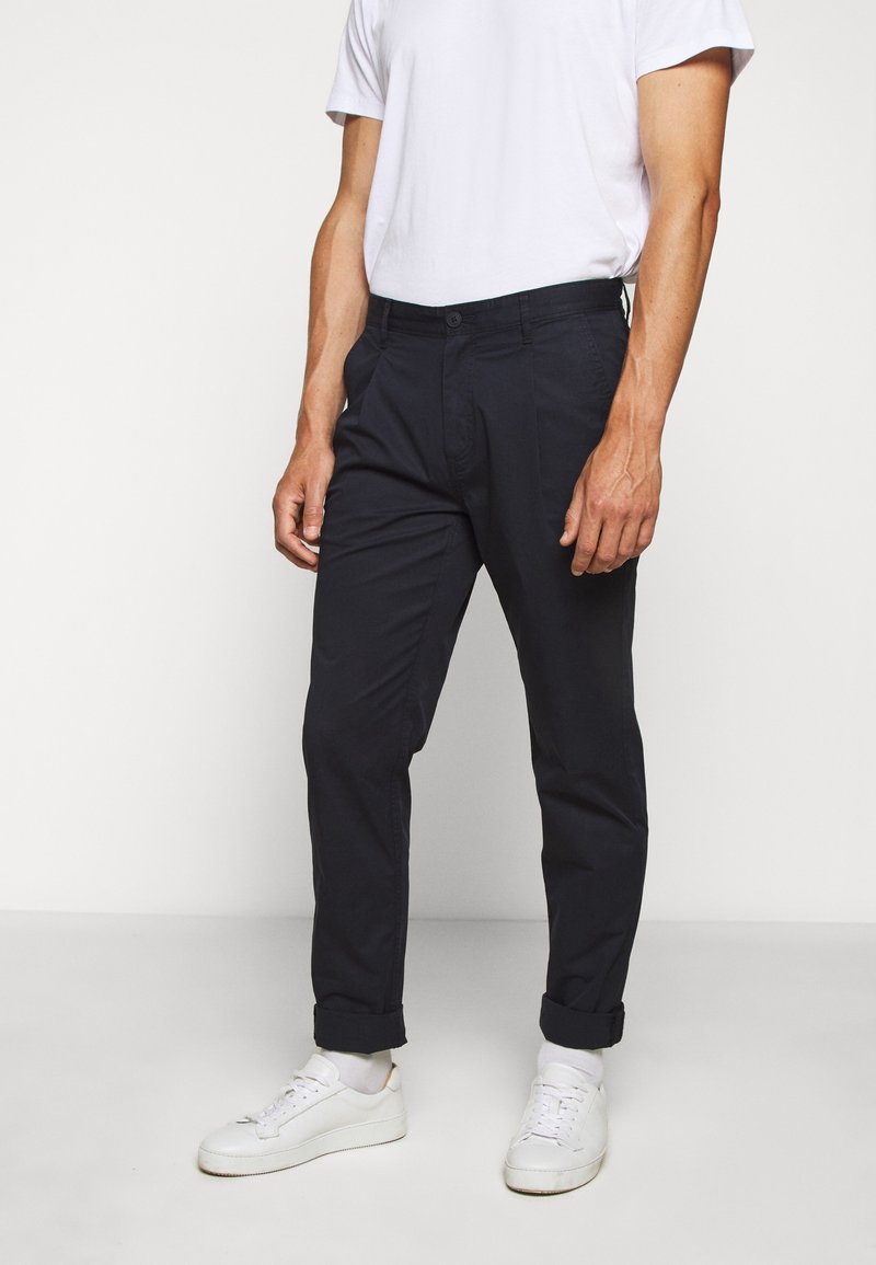 Homme portant un t-shirt blanc à manches courtes, un pantalon sombre retroussé et des baskets blanches, debout devant un fond clair et uni.