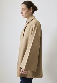 Béžový, veľký top s golierom, dlhými zvonovými rukávmi a zapínaním na gombíky. Hladký materiál, minimalistický dizajn, kombinovaný s modrými džínsami.