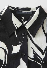 Camicia a maniche lunghe bianca e nera con motivo a stampa, colletto classico, due bottoni neri e un tessuto morbido e testurizzato.