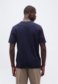 Άνδρας που φοράει μπλε σκούρο κοντομάνικο μπλουζάκι και μπεζ παντελόνι, φαίνεται από πίσω με φόντο ένα απλό ανοιχτόχρωμο φόντο.