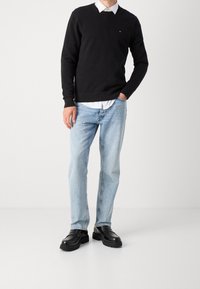 Schwarzer Strickpullover über einem weißen Kragenhemd, hellblaue Jeans und schwarze Schuhe mit einer dicken Sohle. Schlichtes Design, lässiges Outfit.