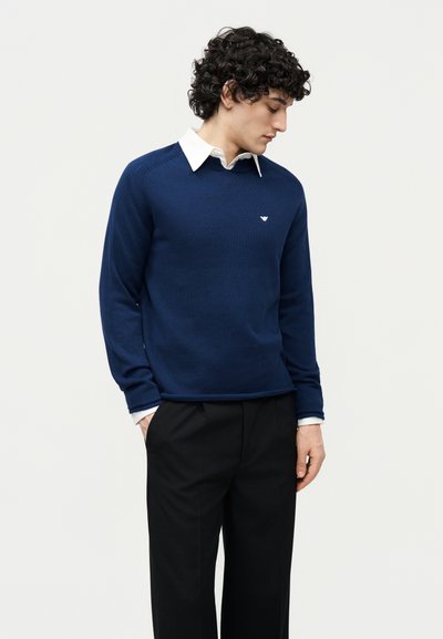 Άνδρας με σγουρά σκουρόχρωμα μαλλιά, που φοράει μπλε σκούρο πουλόβερ πάνω από λευκό πουκάμισο με γιακά και μαύρο παντελόνι, κοιτάζει προς τα κάτω με τα χέρια στις τσέπες.