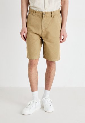 Mann trägt beige knielange Shorts, weiße Socken, weiße Turnschuhe und ein hell gestreiftes Hemd und steht vor einem einfarbigen Hintergrund.