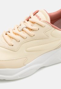 Light peach sneakers with a textured fabric upper, lace-up closure, and a white rubber sole. Features branding on the tongue and contrasting accents.