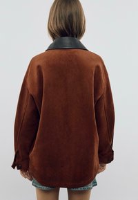 Veste en daim marron avec col en cuir noir. Présente une coupe décontractée et des détails de boutons aux manches, vue de dos.