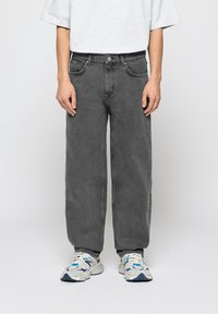 Jean en denim gris à coupe large, avec un design classique à cinq poches et une fermeture par bouton. Porté avec des chaussures de sport blanches et bleues.