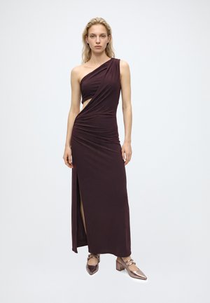 Un mannequin blonde se tient devant un fond de studio clair, vêtue d’une robe de soirée prune foncé à une épaule dénudée, fendue jusqu’à la cuisse, et de chaussures à talons métallisés.
