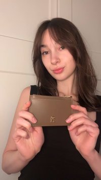 Καφέ δερμάτινη θήκη για κάρτες με χρυσαφένιες λεπτομέρειες, λογότυπο χαραγμένο στην πρόσοψη, λεπτό ορθογώνιο σχήμα, λεία υφή, κρατείται στο χέρι.