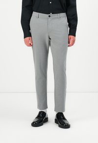 Homme portant un pantalon gris clair sur mesure, une chemise noire à boutons, des chaussettes blanches et des chaussures de ville en cuir noir, debout sur un sol blanc.