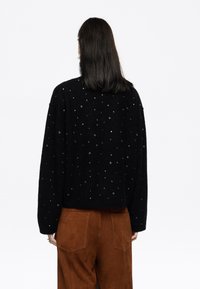 Črn, oversized pulover s srebrnimi okrasnimi elementi. Mehek material, spuščena ramena in sproščen kroj. V kombinaciji z rjavimi suede hlačami.