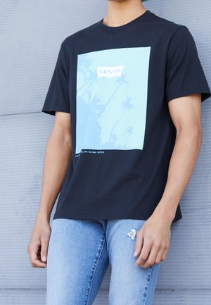 Camiseta de algodón negra con un gráfico azul claro de palmeras y el logo de "Levi's", combinada con unos vaqueros rasgados azul claro.