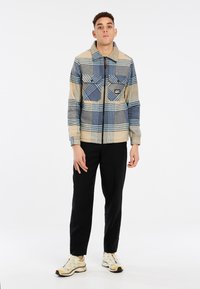 Veste en polaire à carreaux dans des tons beige, bleu et gris ; dotée de deux poches avant et d'un col ; portée avec un pantalon noir et des baskets.