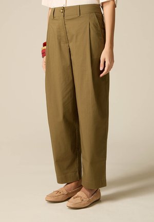 Persona che indossa pantaloni larghi verde oliva, mocassini beige e un top corto bianco, in piedi davanti a uno sfondo semplice.