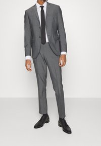 Szary garnitur na miarę o wąskim kroju, zapinana marynarka na guziki, spodnie z płaskim przodem oraz czarny krawat. Całość dopełniają czarne eleganckie buty.