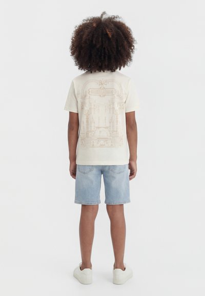 Enfant aux cheveux bouclés debout de dos, portant un t-shirt crème à motif graphique, un short en jean clair et des baskets blanches sur fond blanc.