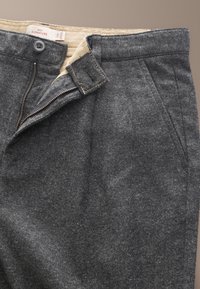 Pantalon en laine gris foncé avec fermeture éclair partiellement ouverte, doublure intérieure beige, fermeture par bouton et poches latérales avant visibles.