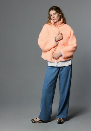 Peach fleece jacket with high collar and blue accents, paired with loose blue pants and dark shoes. Model poses against a gray background.