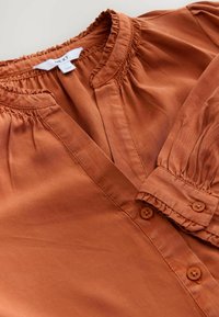 Hedvábná blůza v barvě rzi, která se vyznačuje měkkou tkaninou, nařaseným výstřihem, knoflíkovou légou a nabíranými rukávy s elastickými manžetami.