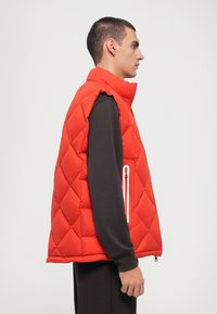 Rød puffervest med diamantquiltet design, høj krave og lynlåslomme foran. Båret over en mørk langærmet skjorte.