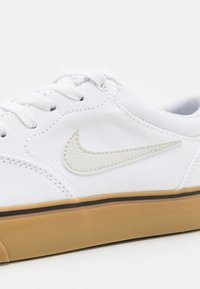 Sapatilha branca de lona com uma superf�ície texturada, sola de borracha bege e logótipo da Nike em tom de bege claro. Apresenta seis ilhós e atacadores brancos.