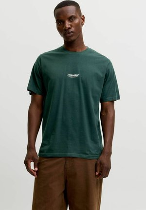 Jeune homme à la peau foncée et aux cheveux courts portant un t-shirt vert JJ Studios et un pantalon marron sur un fond clair uni.