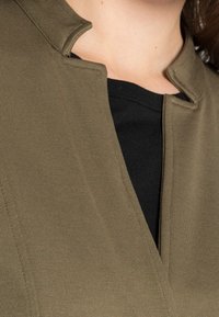 Olive green garment with a mandarin collar, textured fabric, and V-neck design layered over a black top. Close-up, focusing on neckline details.