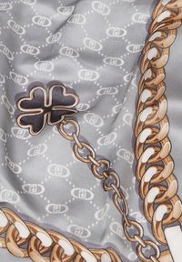Pañuelo de seda con un fondo gris, patrón de cadenas entrelazadas en marrón y dorado, con un motivo de trébol de cuatro hojas contorneado en negro.