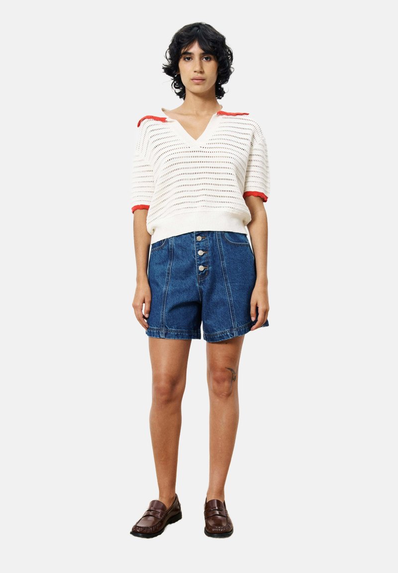 Weiße Strickoberteil mit roten Akzenten, kombiniert mit blauen Jeansshorts mit Knopfverschluss. Das Model trägt braune Slip-On-Schuhe und steht in Pose.