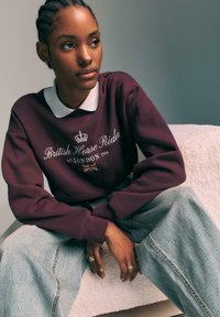 Bordo pulover s belim ovratnikom, napisom "Britanski konjeniški dreser" in dizajnom krone. V kombinaciji z svetloblušči širokimi hlačami. Tekstura ozadja.