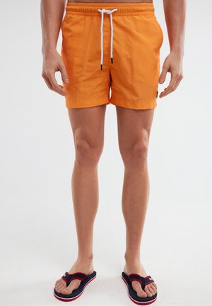 Orange Badehosen mit elastischem Bund und weißem Kordelzug. Kombiniert mit marineblauen Flip-Flops mit roten und weißen Akzenten.