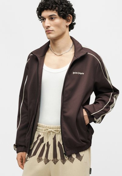 Νέος άνδρας που φοράει καφέ μπουφάν με φερμουάρ και λευκή ρίγα, λευκό φανελάκι, μπεζ παντελόνια με σκούρο γεωμετρικό σχέδιο και ασημένιο κολιέ με αλυσίδα.