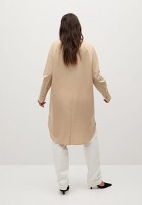 Chemise beige à manches longues avec une coupe droite, poignets boutonnés et une couture légèrement arrondie, associée à un pantalon blanc et des chaussures noires.