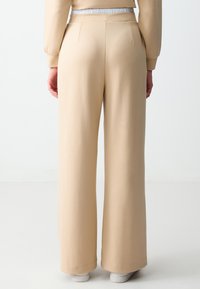Beige wijde broek met een gladde textuur, voorzien van een platte tailleband en subtiele achterzakken. De zoomafwerking is netjes en elegant.