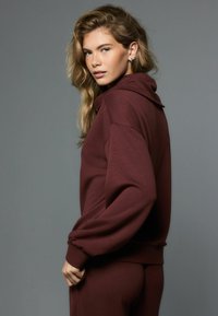 Bordo spalvos džemperis su gaubtu iš minkštos medžiagos, laisvo silueto, su gaubtu ir ribinėmis rankogaliais. Džemperis derinamas su atitinkančiomis apačiomis.