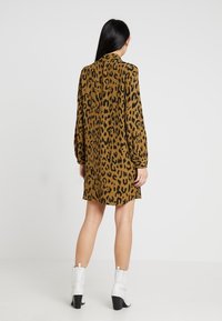 Abito marrone con stampa leopardata, maniche lunghe, colletto e silhouette dritta. Il modello indossa stivaletti bianchi con tacco nero.