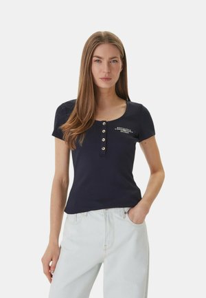 Giovane donna con capelli castani lunghi, indossa un top nero a maniche corte con bottoni e jeans azzurri, in piedi con una mano in tasca.