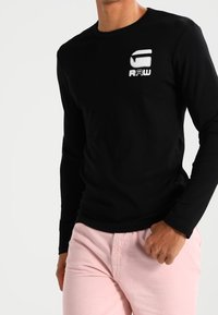 Man met een zwart T-shirt met lange mouwen en een wit G RAW-logo op de borst, in lichtroze broeken, poseert tegen een witte achtergrond.